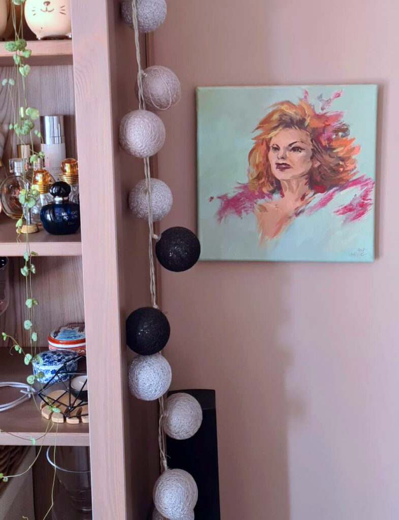 Na różowej ścianie portret kobiety. Wyłania się z błękitu. Ma wielokolorowe włosy, głównie różowe oraz pomarańczowe. Ma skwaszoną minę. Na lewo od pracy szafka oraz lampki.