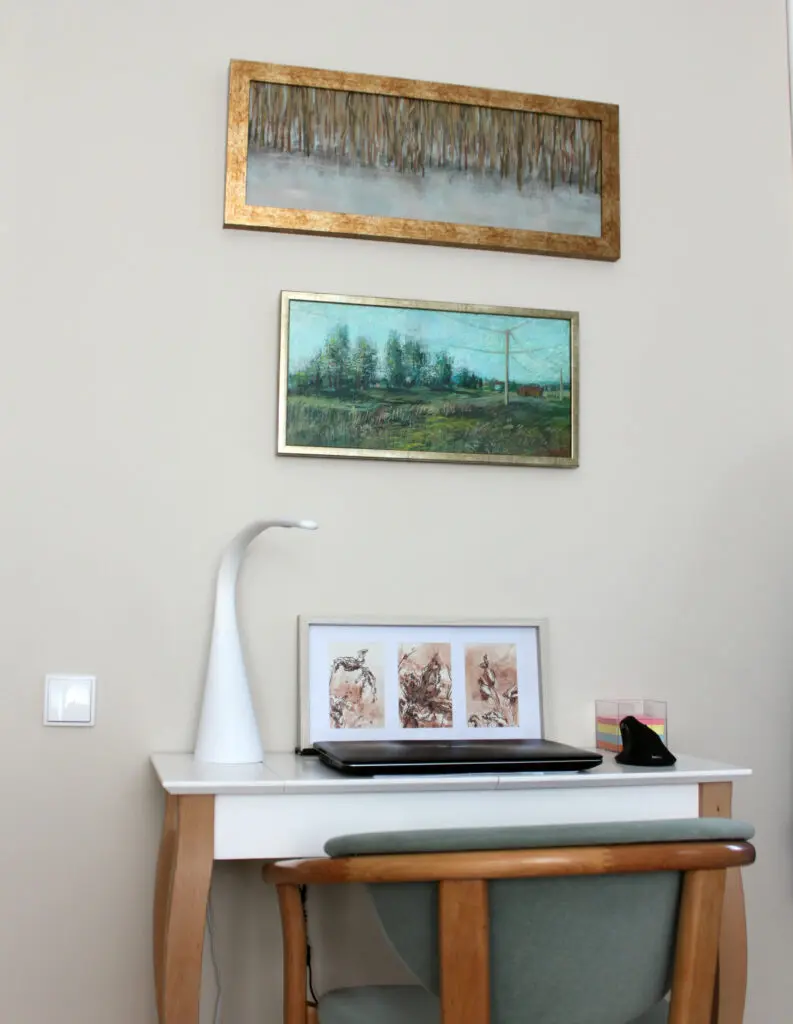 Biała ściana oraz biurko. Na biurku obraz złożony z trzech pomniejszych, brązowych grafik. Na ścianie dwa obrazy. Górny przedstawia jezioro oraz szuwary. Na dolnym sielankowy krajobraz wsi.