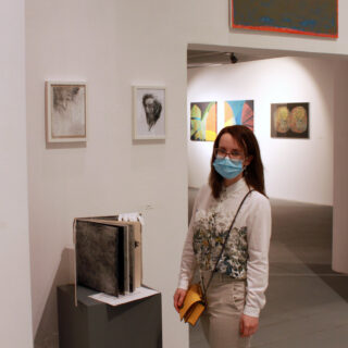 Młoda artystka na wernisażu wystawy przy swoich pracach. Ubrana w koszulę w kwiaty oraz szare spodnie. Ma żółtą torebkę. Nad nią jej obrazy.