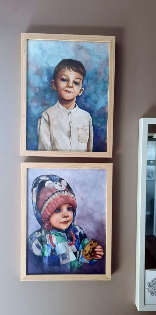 Dwa portrety w złotych ramach, jeden nad drugim. Górny przedstawia małego chłopca w białej koszuli na niebieskim tle. Na dolnym mała dziewczynka w zimowej czapce oraz kurtce, w dłoni trzyma ciastko.