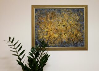 Na rośliną na ścianie w złotej ramie abstrakcyjna praca. Jest złoto-granatowa. Może przypominać rój pszczół na tle plastrów miodu.