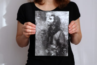Młoda kobieta trzyma obraz stylizowany na starą kartę. Na nim mężczyzna z bujnym zarostem, ubrany w koszulę i sweterek. Pali papierosa. W rogach pracy symbol króla karcianego.